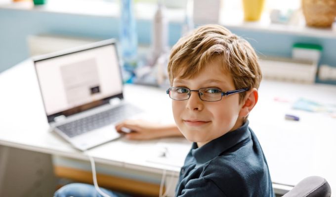 chłopiec w okularach siedzący przy biurku na którym stoi otwarty laptop.
