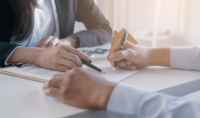 obrazek pokazuje zbliżenie na ręce kobiety i mężczyzny, kobieta wskazuje miejsce do podpisu w dokumencie leżącym na biurku